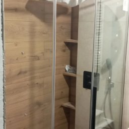 Zakład Produkcyjno - Handlowy MIKOR Andrzej Wudzki - Nowoczesna kabina prysznicowa z drewnopodobną okładziną ścienną i półkami wewnątrz. Minimalistyczny design, czarne detale armatury, odpływ liniowy w podłodze.