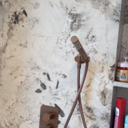 Zakład Produkcyjno - Handlowy MIKOR Andrzej Wudzki - Nowoczesna bateria prysznicowa w odcieniach brązu na tle ściany z efektem marmuru. Widoczne kosmetyki na półce. Minimalistyczny design.