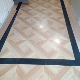 Zakład Produkcyjno - Handlowy MIKOR Andrzej Wudzki - Ukończona podłoga z wzorem parkietu w jasnym odcieniu drewna, obramowana czarnymi płytkami. Część paneli jeszcze nieułożona, leżą obok ściany.