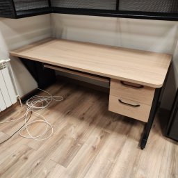 Jakub Dera USŁUGI OGÓLNOBUDOWLANE - Drewniany biurko z metalowymi nogami i szufladami, widok z góry, na podłodze leżą białe kable. Nad biurkiem metalowa półka z siatki.