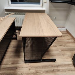 Jakub Dera USŁUGI OGÓLNOBUDOWLANE - Stół z drewnianym blatem i metalową, ażurową podstawą w industrialnym stylu. Widoczne fragmenty drugiego stołu i podłoga z paneli.