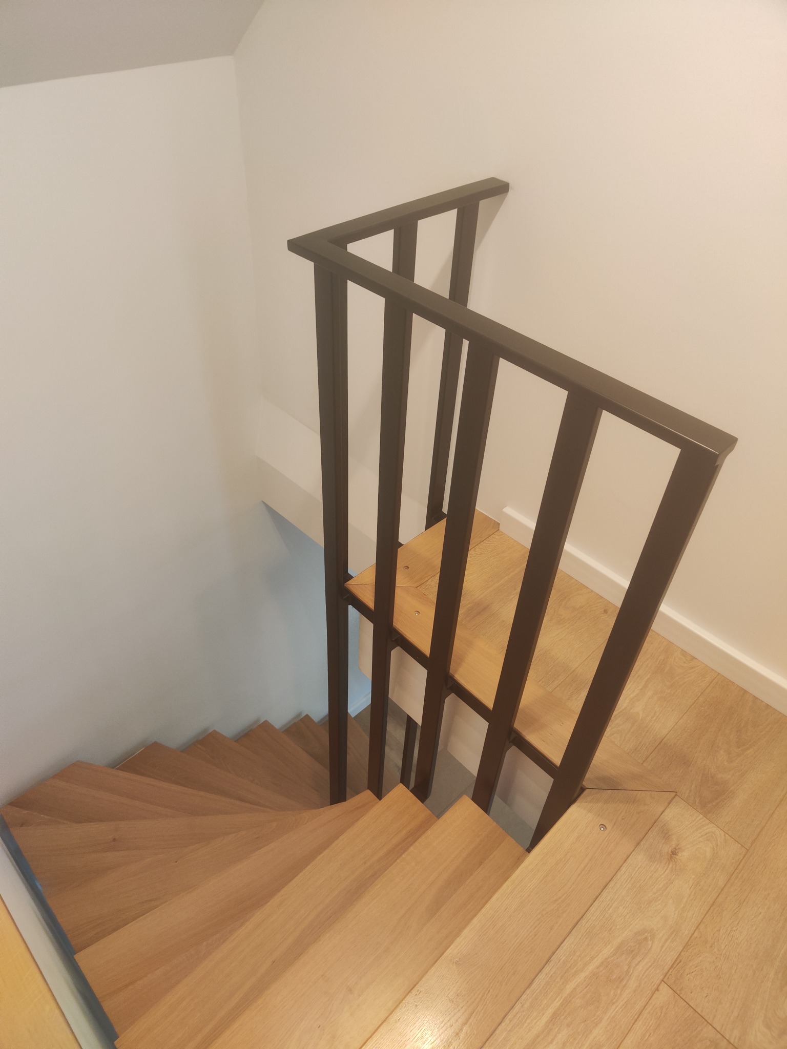 Drewniane schody z czarną, metalową balustradą, widziane z góry. Naturalne oświetlenie podkreśla strukturę drewna i minimalistyczny design. Nowoczesne wykończenie wnętrza.