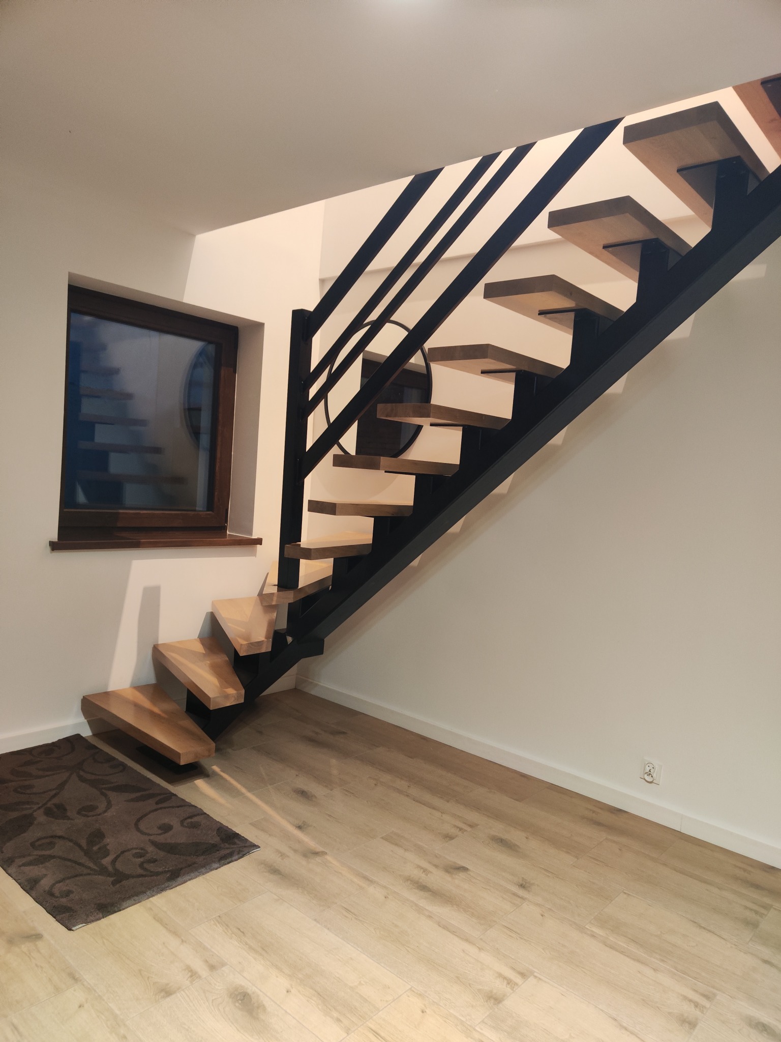 Nowoczesne schody metalowe z drewnianymi stopniami w jasnym wnętrzu. Minimalistyczny design, czarna konstrukcja, drewno w ciepłym odcieniu. Widok z dołu, dywanik przy schodach.