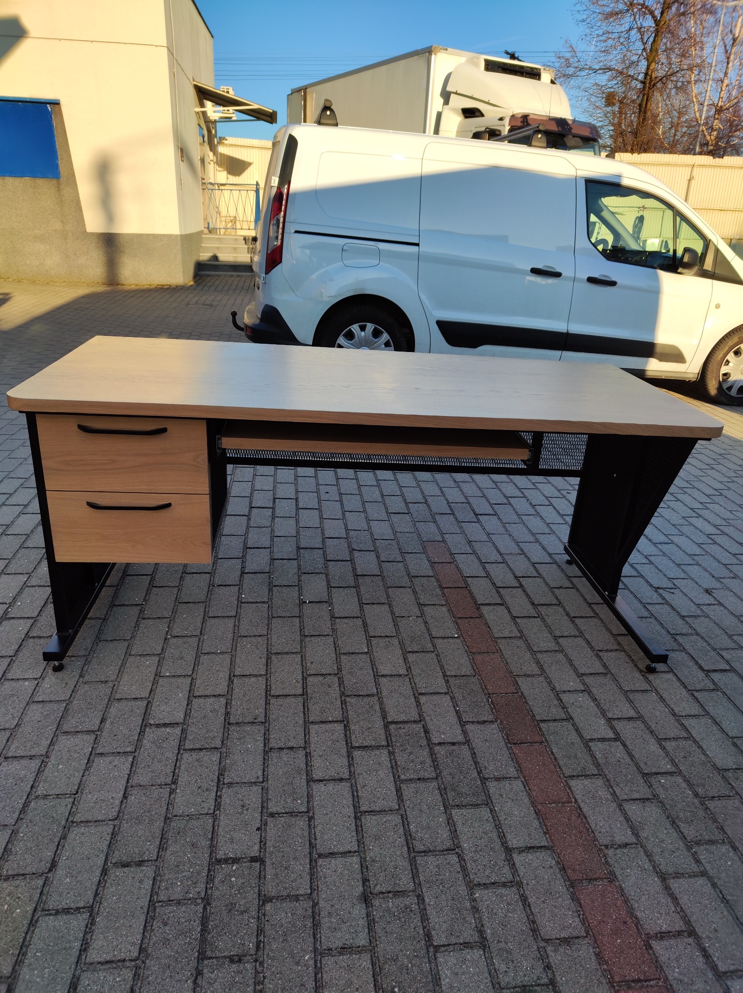 Stół biurowy z drewnianym blatem i czarną metalową konstrukcją na zewnątrz, z białą furgonetką w tle, prezentowany na brukowanej powierzchni.