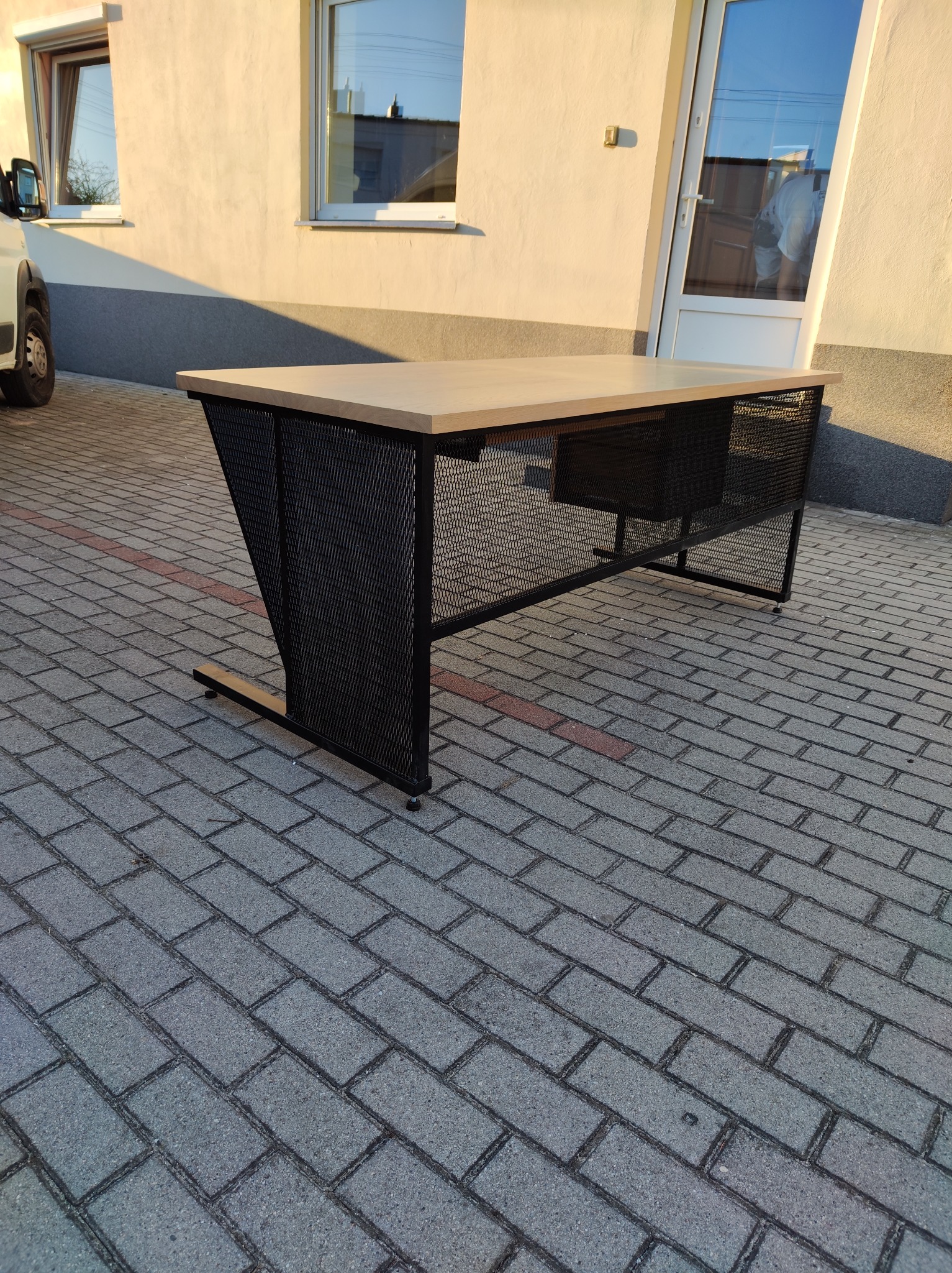 Nowoczesne biurko z blatem w kolorze drewna i metalową, ażurową konstrukcją, ustawione na brukowanym podjeździe przed budynkiem. Widoczne okna i drzwi.