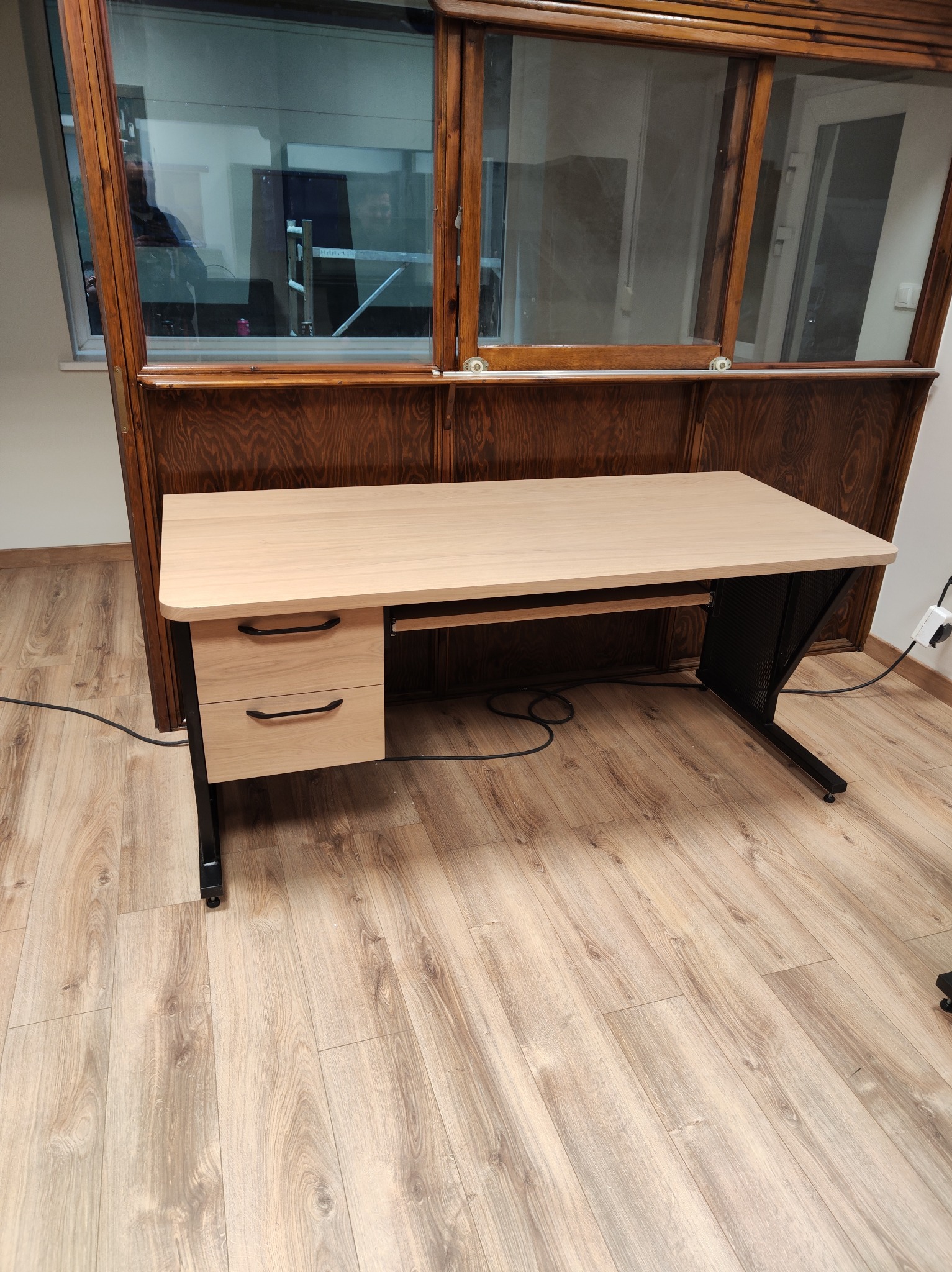 Biurko z jasnego drewna z czarnymi nogami i szufladami, ustawione na panelach podłogowych. W tle widoczna drewniana zabudowa okienna.