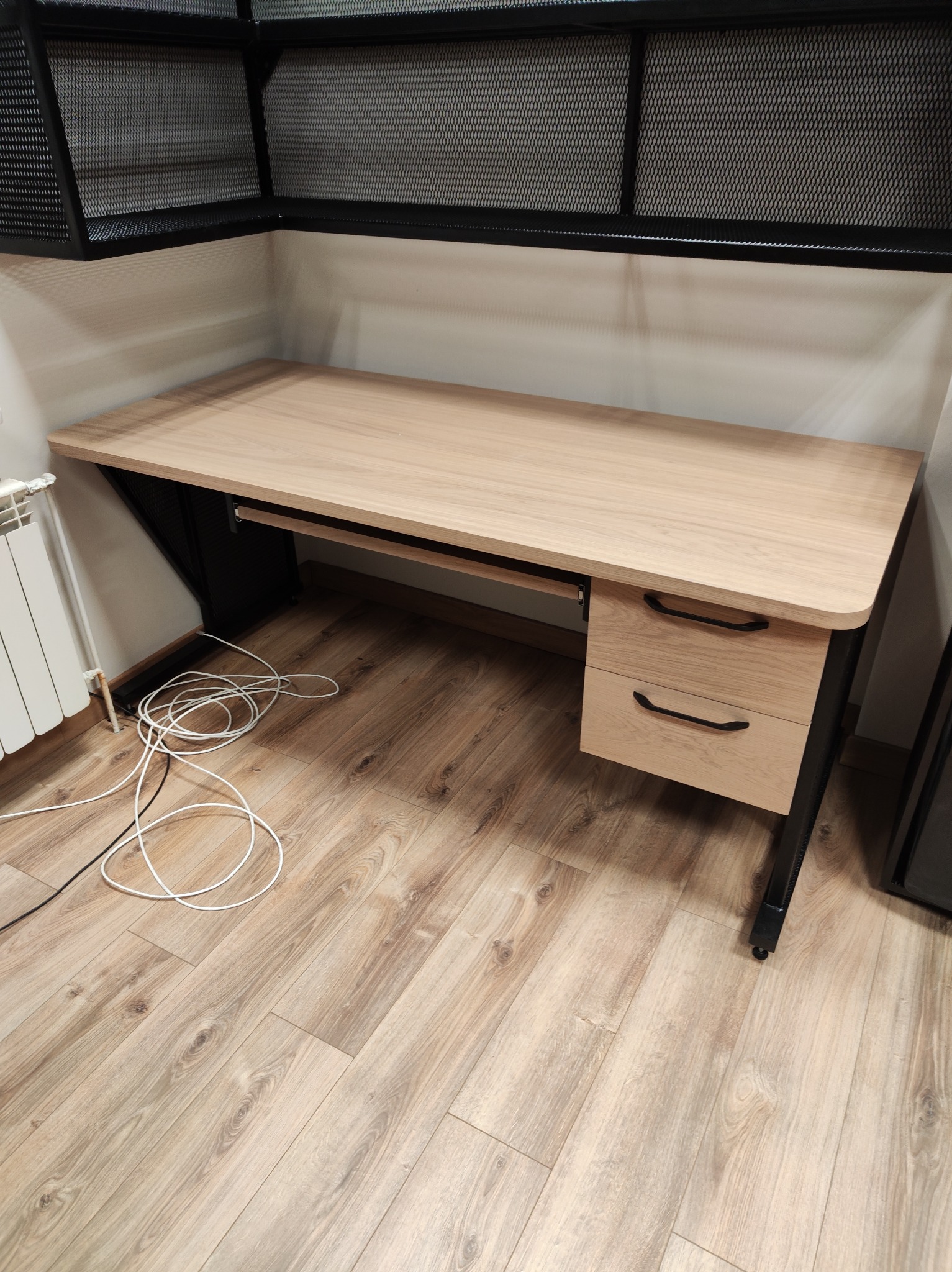 Drewniany biurko z metalowymi nogami i szufladami, widok z góry, na podłodze leżą białe kable. Nad biurkiem metalowa półka z siatki.