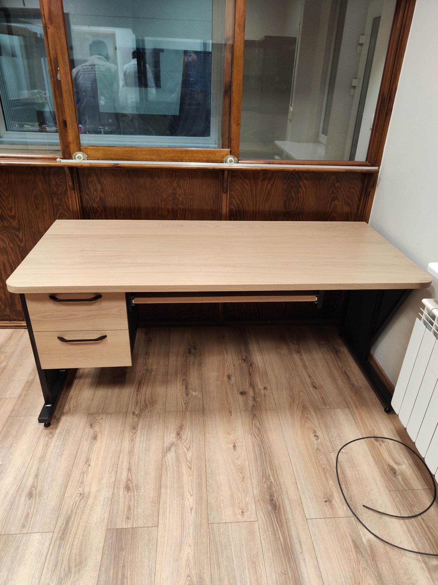 Biurko z jasnego drewna z czarnymi nogami i szufladami, ustawione na drewnianej podłodze w pomieszczeniu z oknem. Widoczny grzejnik i kabel na podłodze.