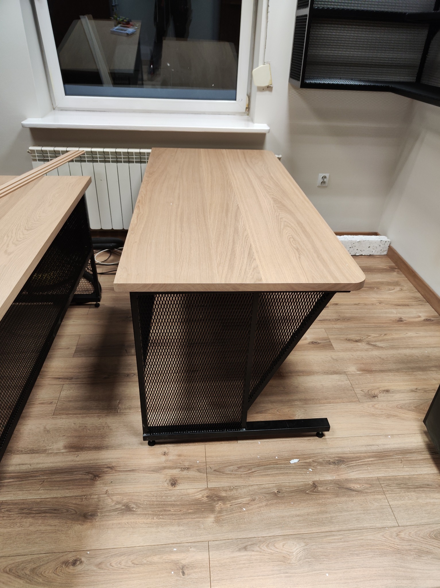 Stół z drewnianym blatem i metalową, ażurową podstawą w industrialnym stylu. Widoczne fragmenty drugiego stołu i podłoga z paneli.