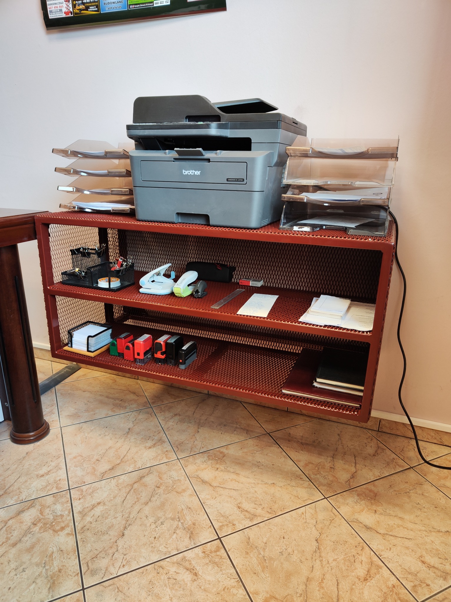 Metalowy regał z siatki w kolorze ceglastym, z drukarką Brother i akcesoriami biurowymi. Widok z przodu, na tle jasnej ściany i podłogi z płytek.