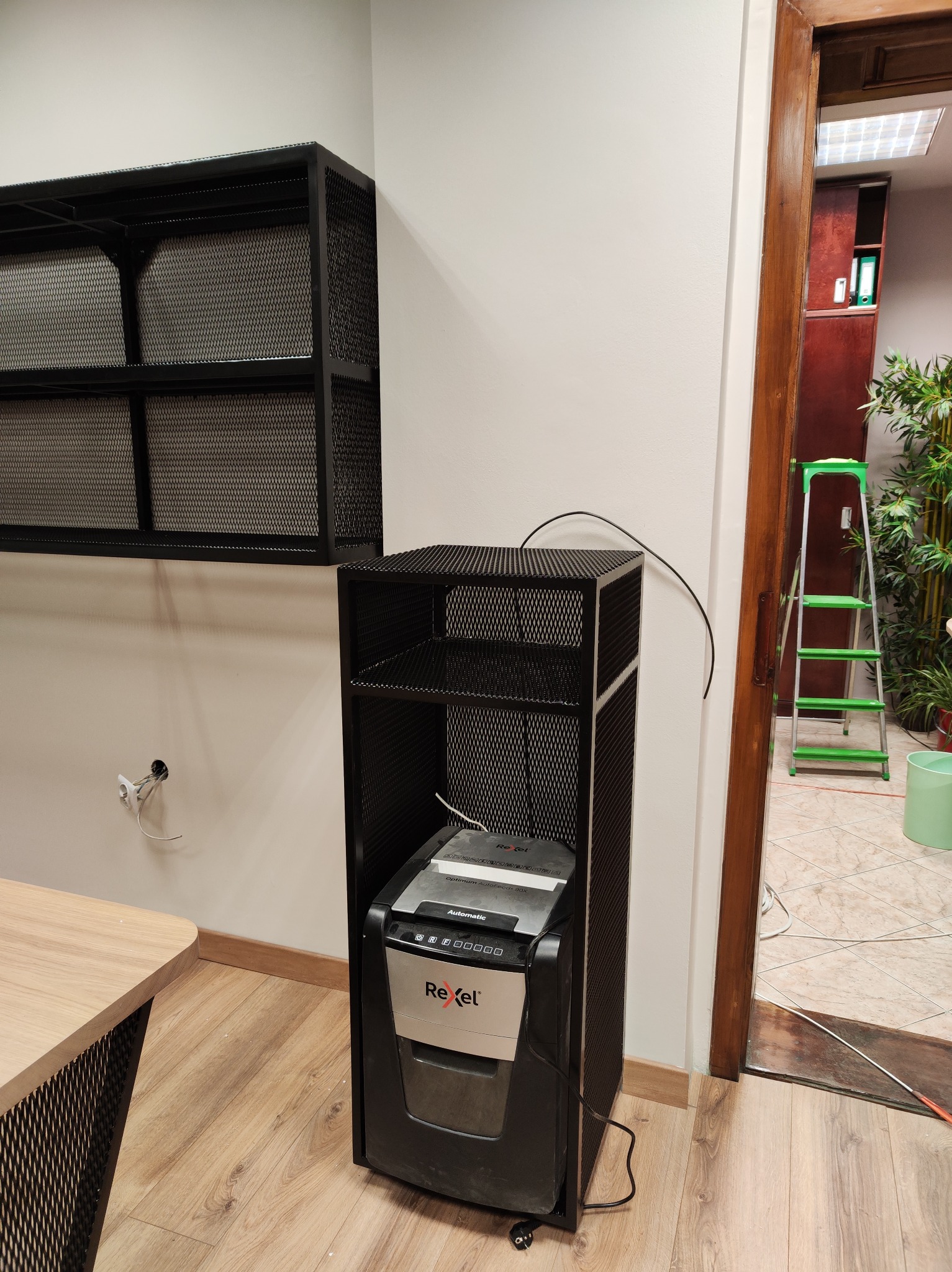 Metalowa półka wisząca i stojąca z perforowanej blachy w biurze. Widoczny fragment biurka z podobnym wykończeniem. W tle drabina i rośliny doniczkowe.