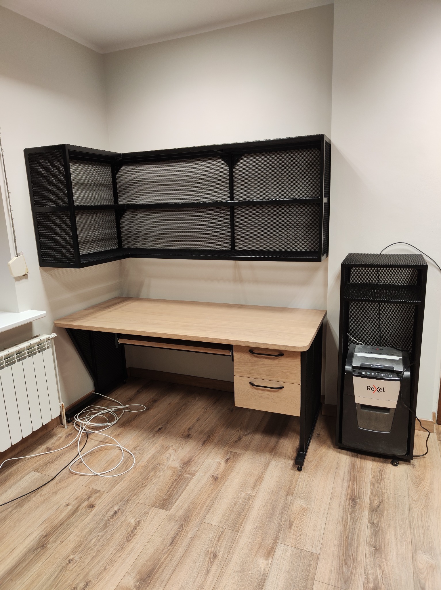 Metalowa konstrukcja biurka z półkami i szafką, obok niszczarka do dokumentów. Całość w stylu industrialnym, na drewnianej podłodze.