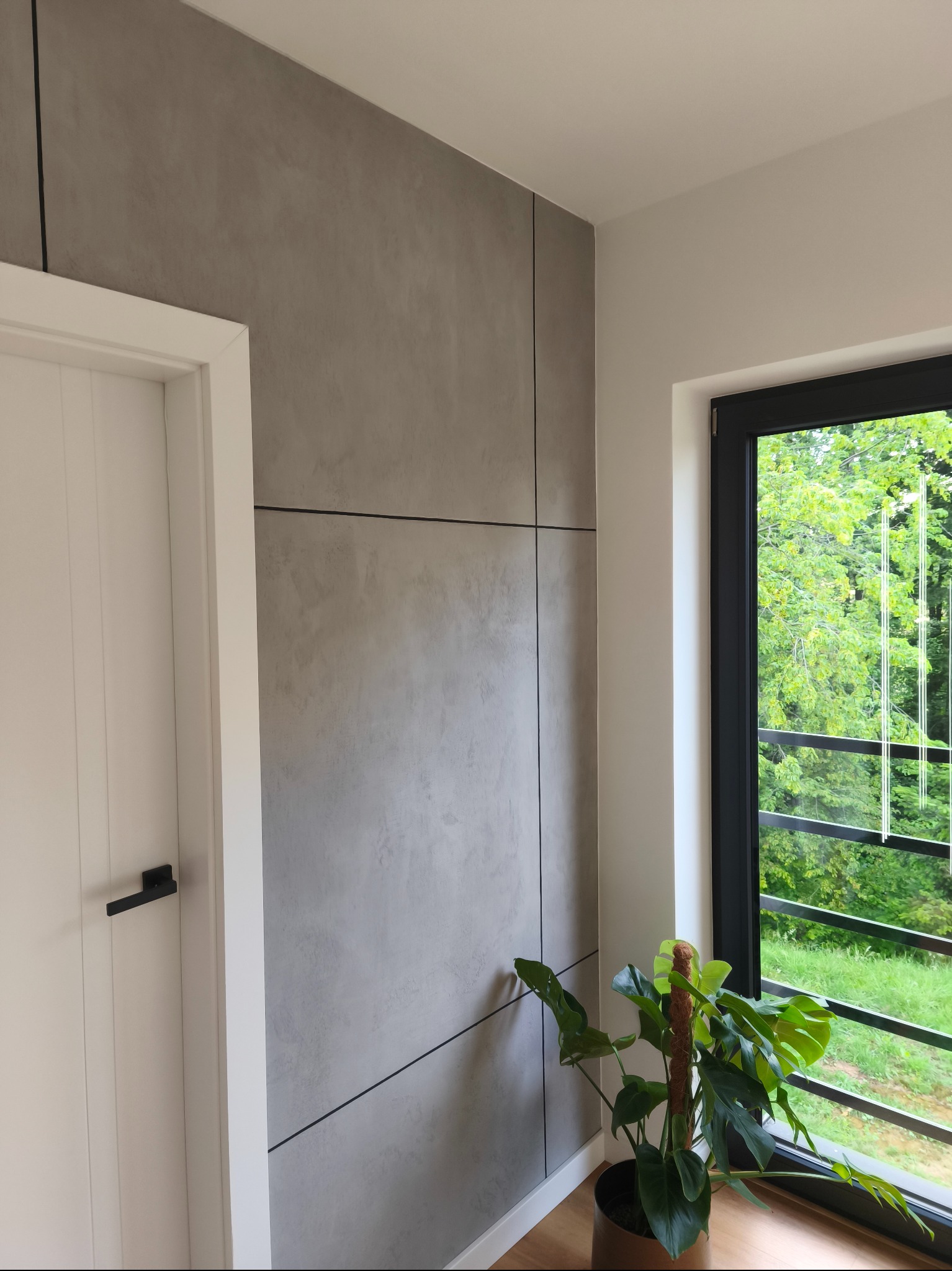 Ściana z szarego betonu architektonicznego podzielona czarnymi listwami, białe drzwi z czarną klamką, okno z widokiem na zieleń, roślina doniczkowa. Nowoczesne wykończenie wnętrza.