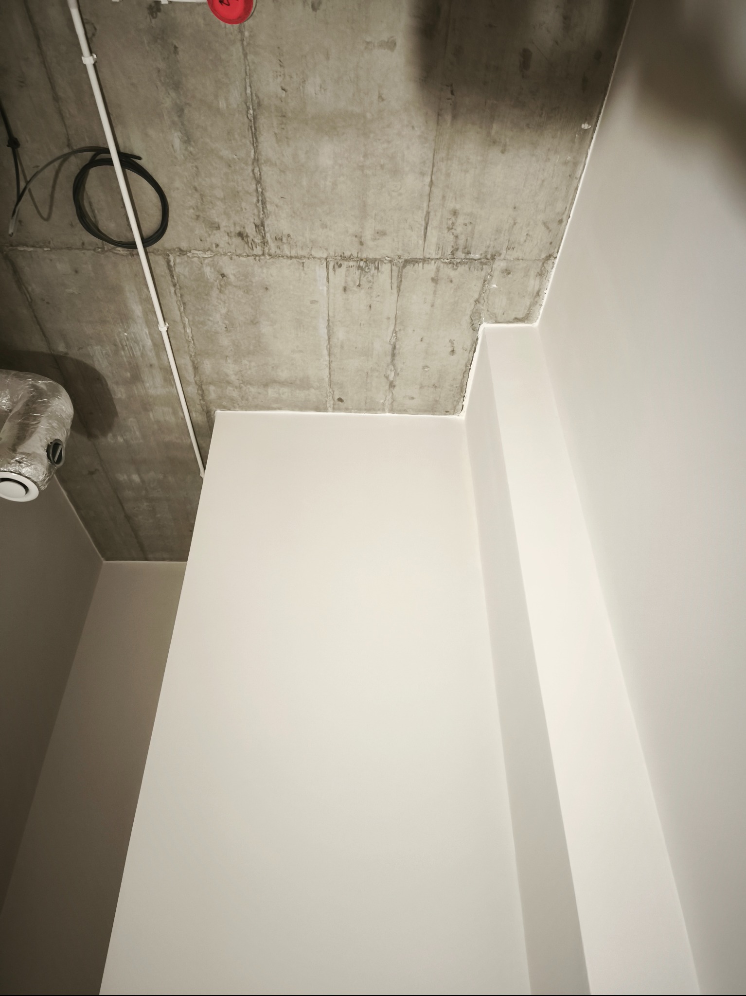 Surowy betonowy sufit z instalacjami i świeżo pomalowane, gładkie, białe ściany w narożniku pomieszczenia. Widoczny fragment wentylacji.