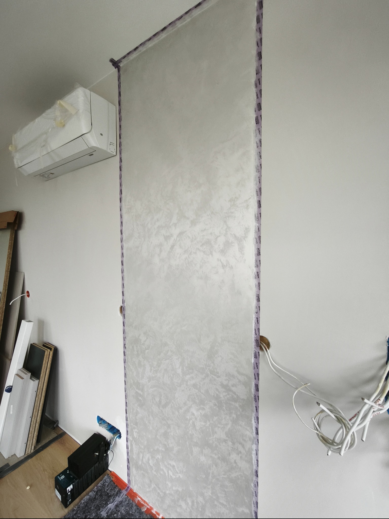 Ściana z dekoracyjną strukturą, zabezpieczona taśmą malarską. Widoczny klimatyzator w folii, kable elektryczne i narzędzia malarskie. Wykończenie wnętrz w trakcie realizacji.