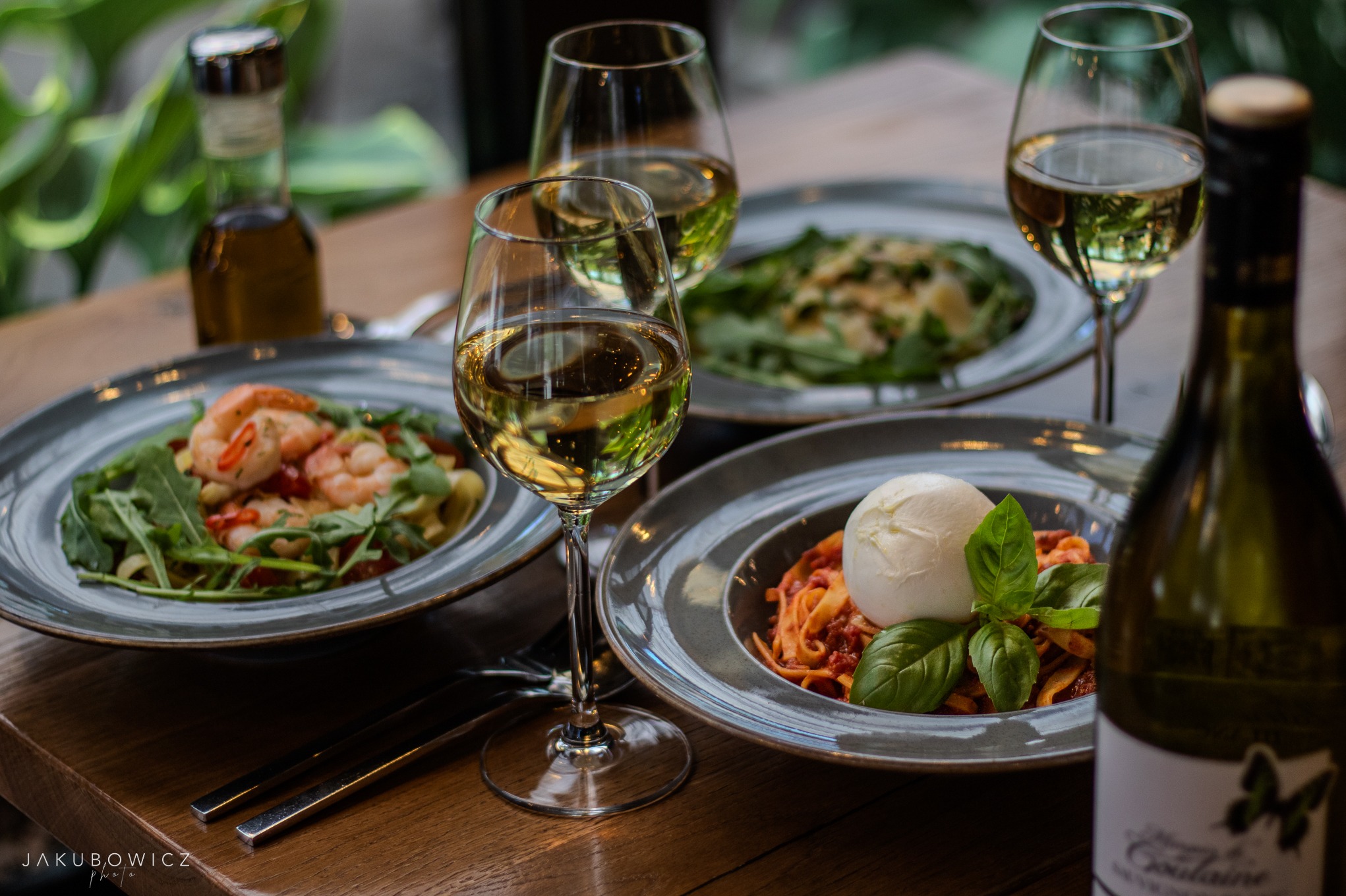 Profesjonalna fotografia produktowa: trzy talerze z daniami (makaron z krewetkami, makaron z burratą i pesto) oraz kieliszki z białym winem na drewnianym stole.