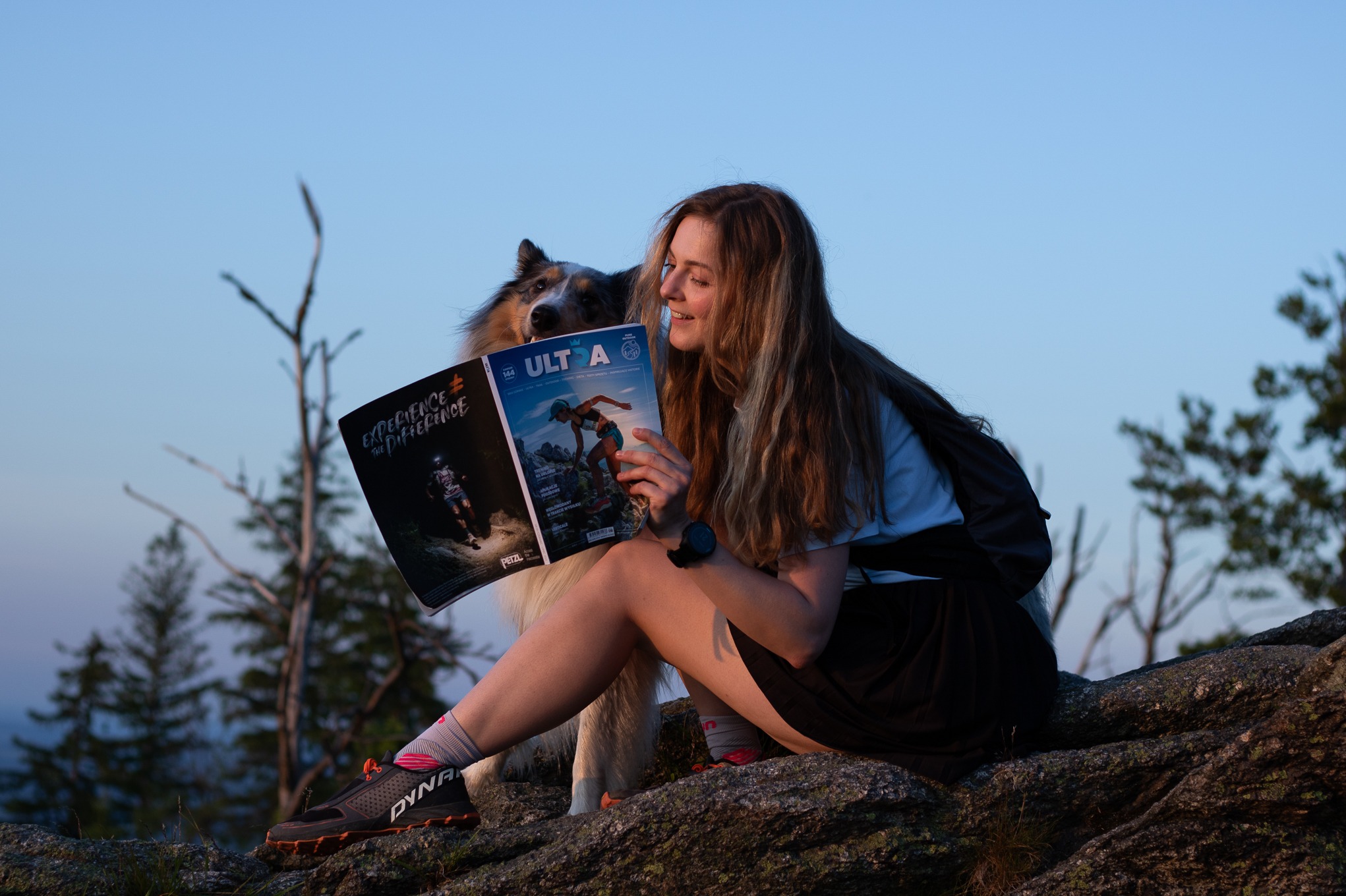 Kobieta z psem siedzi na skale, czytając magazyn o biegach górskich. W tle drzewa i niebo o zachodzie słońca. Widoczne buty do biegania i zegarek sportowy.