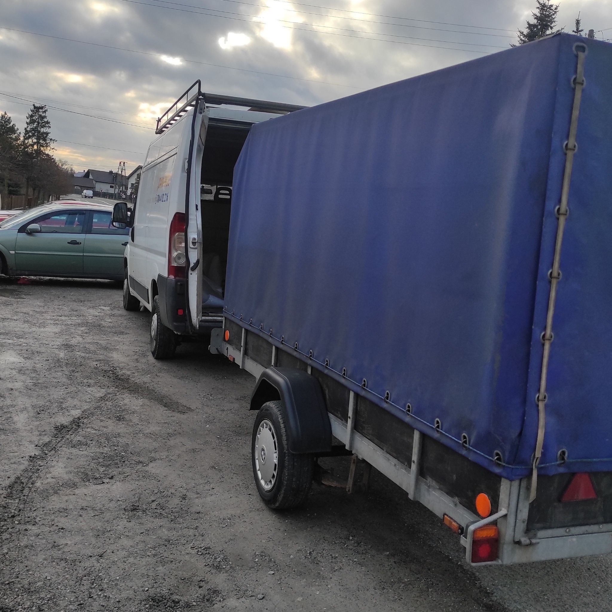 Biały bus z otwartymi drzwiami i podłączoną przyczepą z niebieską plandeką na żwirowym podłożu, w tle zaparkowane samochody i budynki.