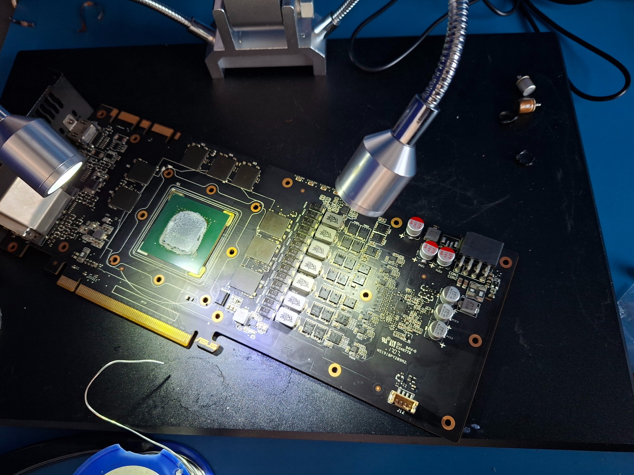 Karta graficzna ASUS po wymianie kondensatorów, oświetlona lampami warsztatowymi. Widoczne ślady lutowania i elementy elektroniczne. Sprzęt IT na czarnym podłożu.