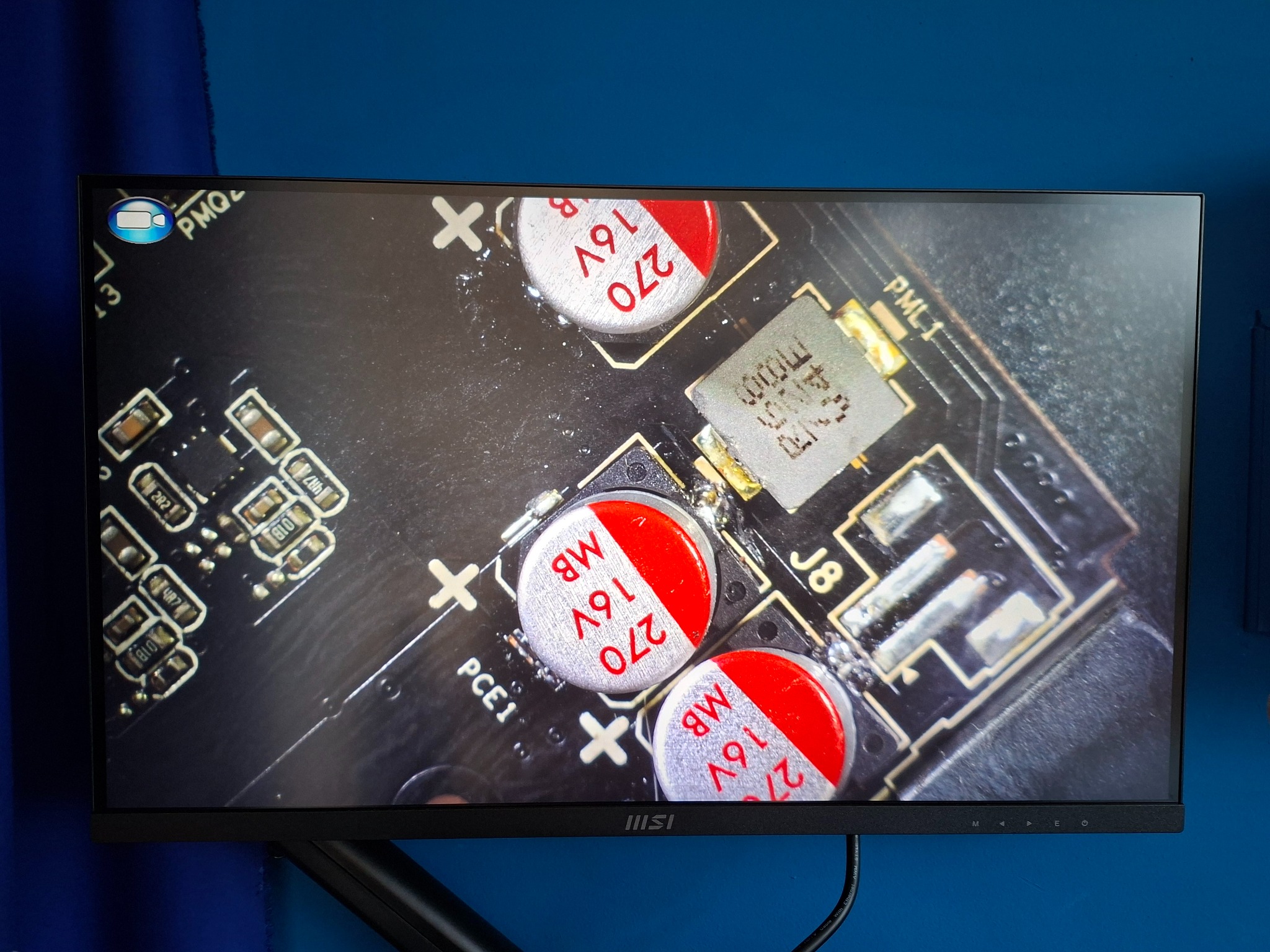 Ekran monitora MSI z powiększonym widokiem płyty głównej komputera, ukazując detale kondensatorów i układów scalonych. Widoczne oznaczenia i elementy lutowane.