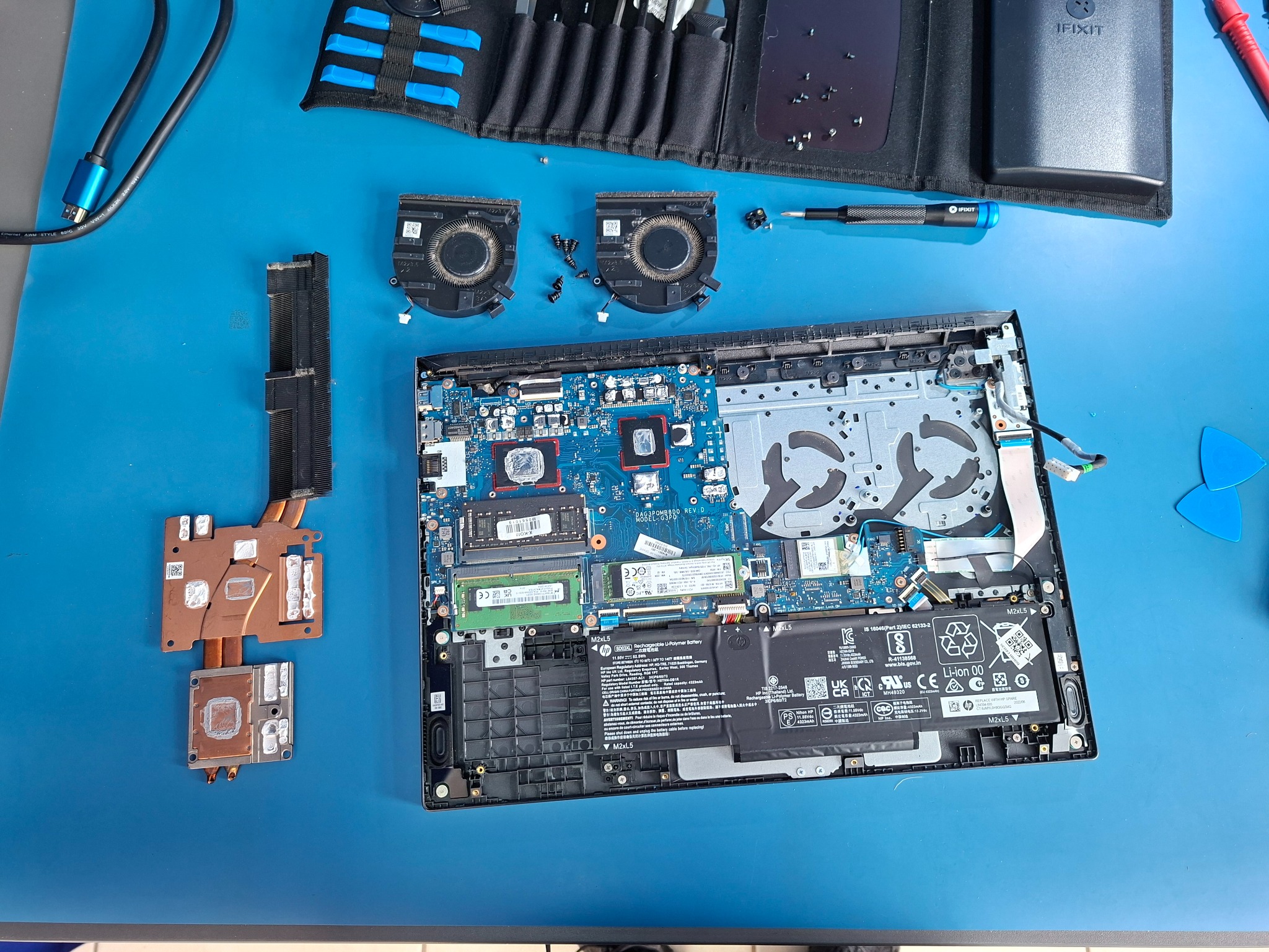 Otwarty laptop z widoczną płytą główną, wentylatorami i elementami chłodzenia, rozłożony na niebieskim podłożu, obok narzędzia i akcesoria do naprawy.
