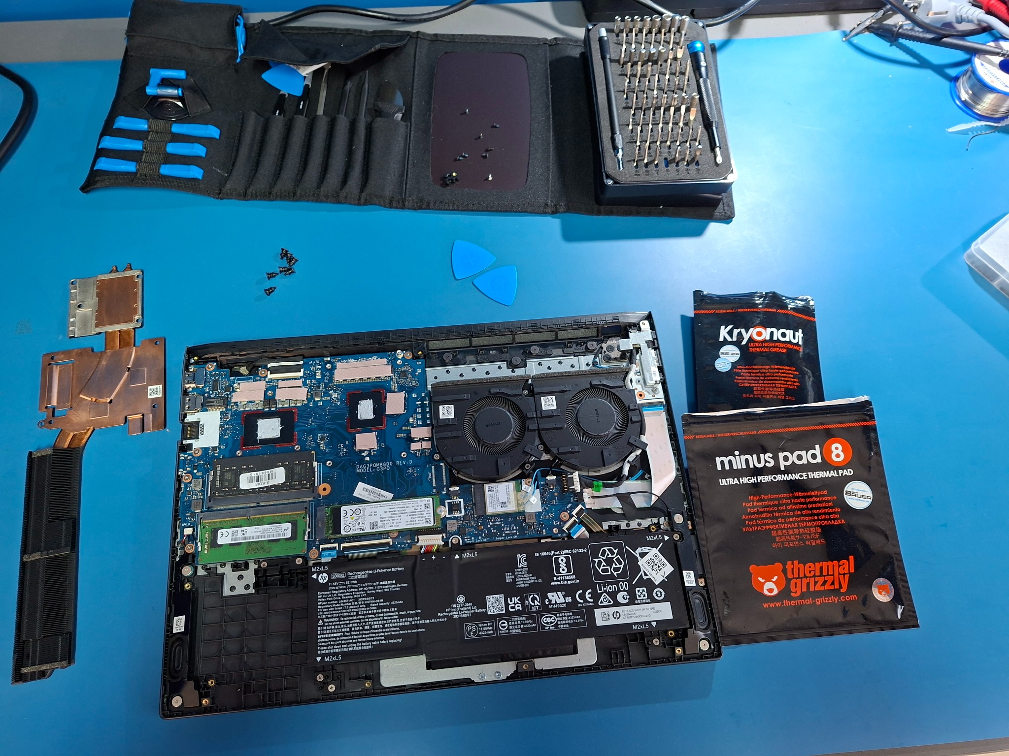 Otwarty laptop z widoczną płytą główną, obok narzędzia, śrubokręty, pasta termoprzewodząca i podkładki termiczne. Widok z góry na stanowisko pracy serwisanta komputerowego.