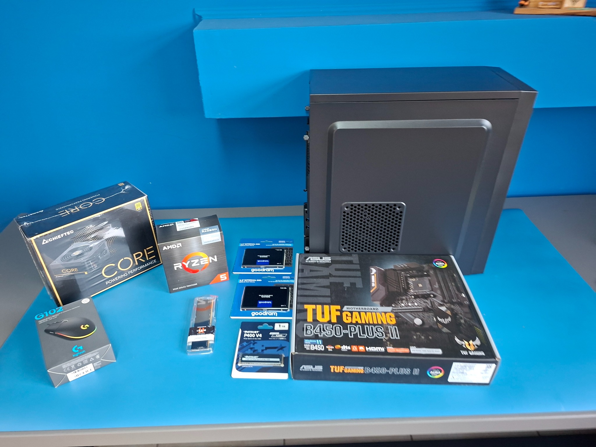 Komponenty PC na niebieskim tle: obudowa, płyta główna ASUS TUF Gaming B450-PLUS II, procesor AMD Ryzen 5, pamięć RAM Goodram, chłodzenie Chieftec Core i mysz Logitech G102.