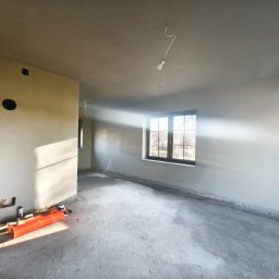 ART-MASTER BARTŁOMIEJ GÓRNIAK TYNKI, DEKORACJE & WYKOŃCZENIA - Surowe wnętrze po tynkowaniu: światło słoneczne wpada przez okno, oświetlając gładkie, szare ściany i betonową podłogę. Widoczne narzędzia i instalacje elektryczne.