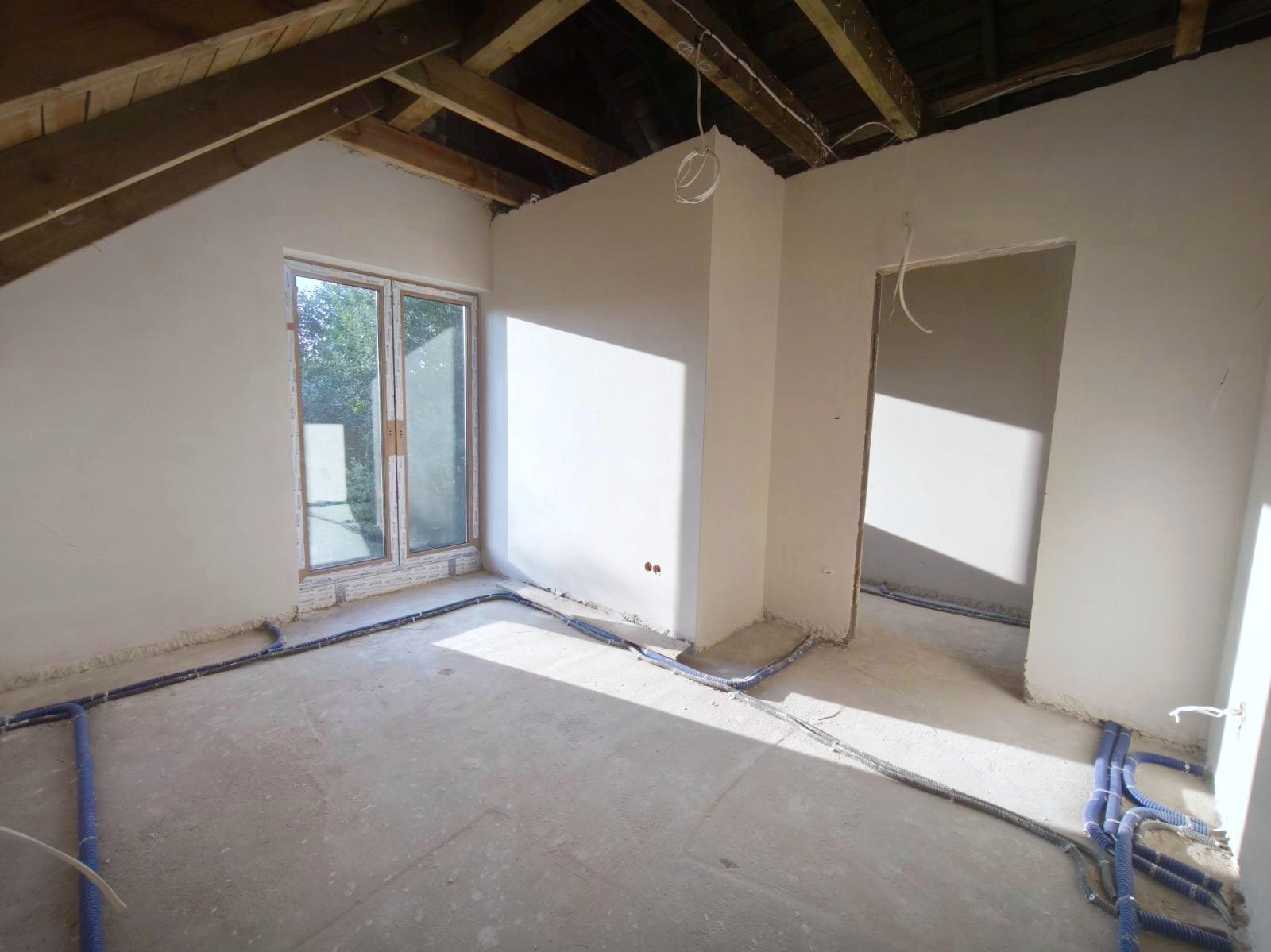 Wnętrze po nałożeniu gładzi w Obornikach Śląskich. Widoczne instalacje elektryczne w podłodze, okno i surowe drewniane elementy konstrukcji dachu.