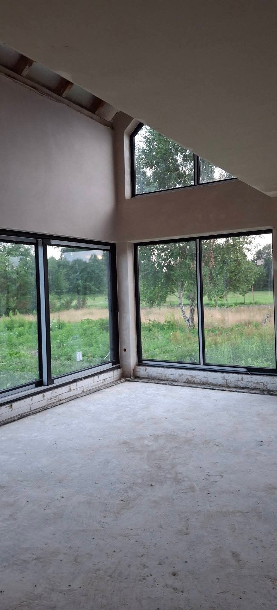 Surowe wnętrze z dużymi oknami i świeżo otynkowanymi ścianami. Widok na zieleń za oknem. Minimalistyczny design, surowy beton na podłodze. Wnętrze w trakcie wykańczania.