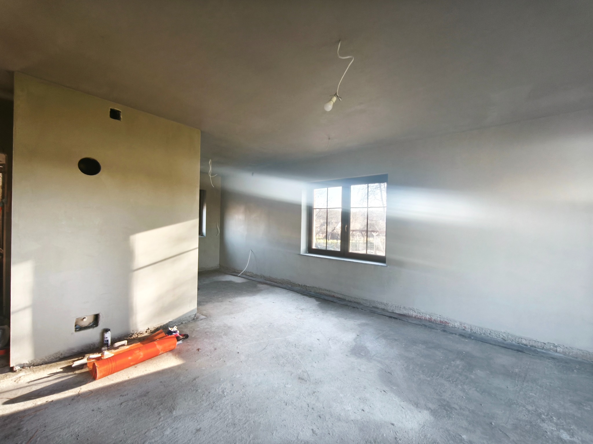 Surowe wnętrze po tynkowaniu: światło słoneczne wpada przez okno, oświetlając gładkie, szare ściany i betonową podłogę. Widoczne narzędzia i instalacje elektryczne.