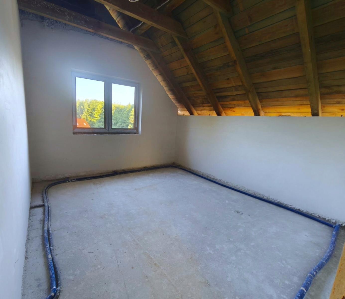 Poddasze z oknem po tynkowaniu ścian, widoczne rury ogrzewania podłogowego przy ścianach i drewniana konstrukcja dachu. Świeże tynki w jasnym odcieniu.