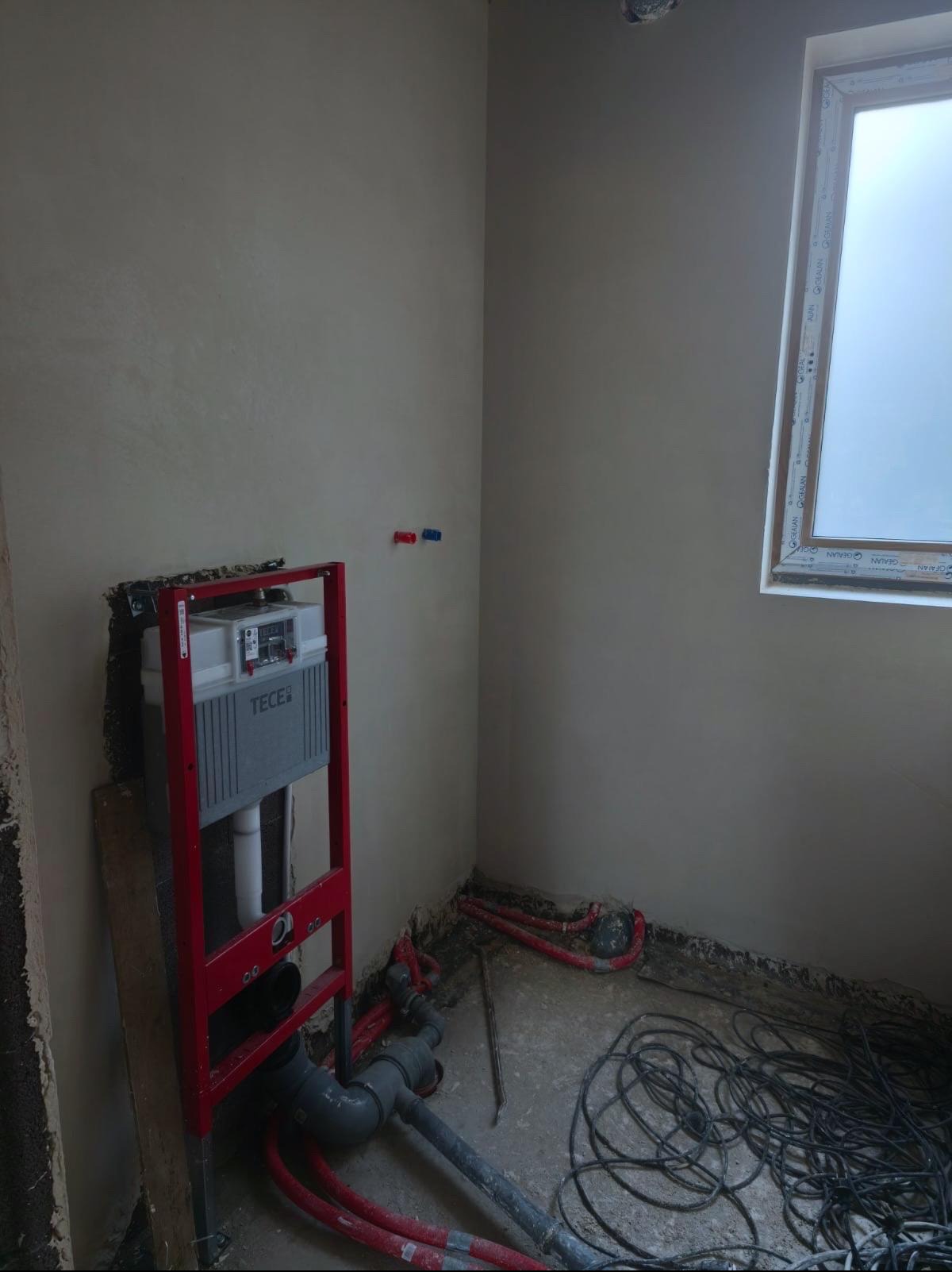 Szara ściana z oknem i instalacją podtynkową WC TECE z czerwonym stelażem. Widoczne rury i kable na podłodze. Ściana pokryta gładzią.