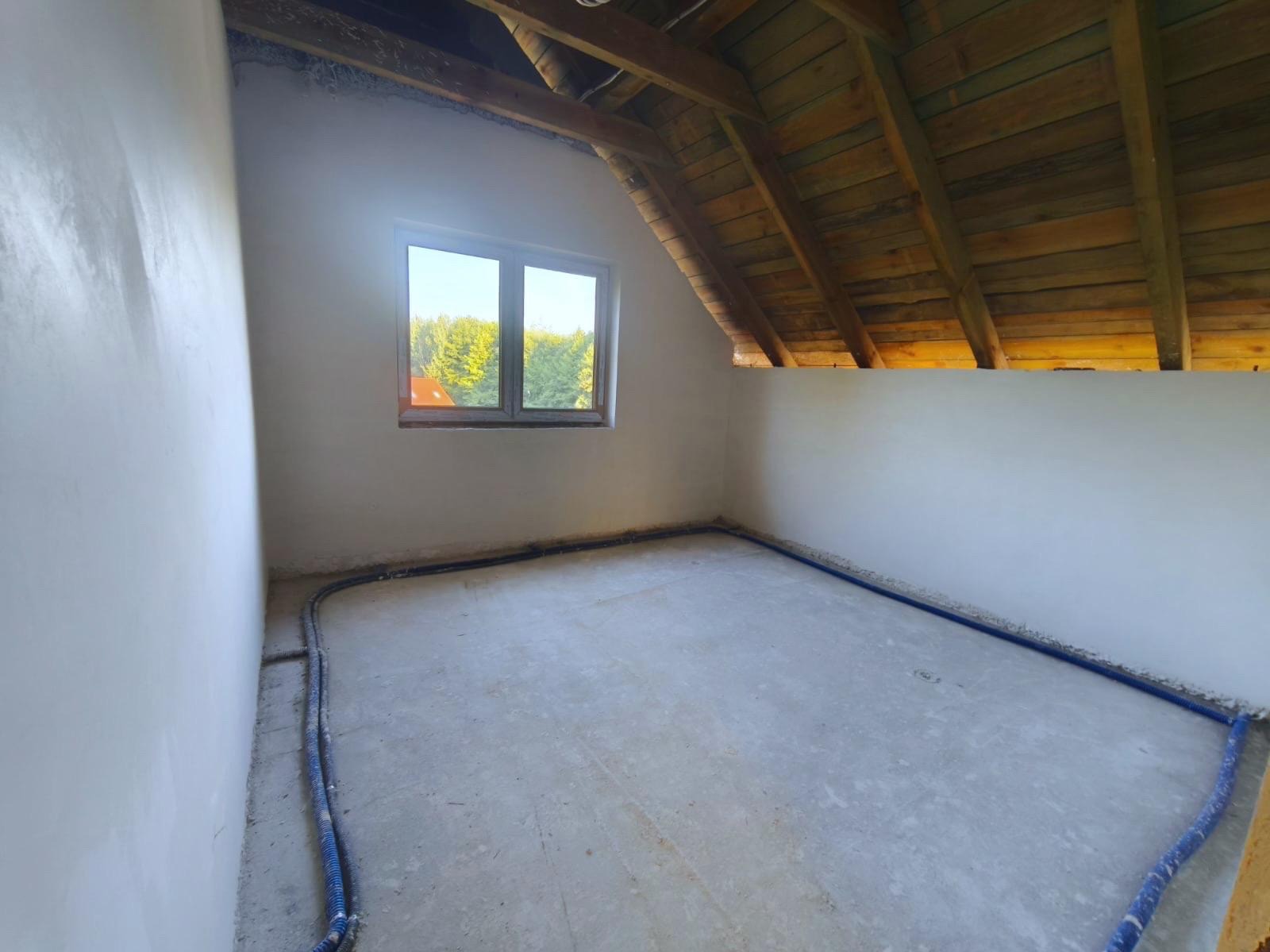 Pokój na poddaszu po tynkowaniu ścian, widoczne drewniane elementy konstrukcji dachu i niebieskie rurki instalacji grzewczej wzdłuż ścian.