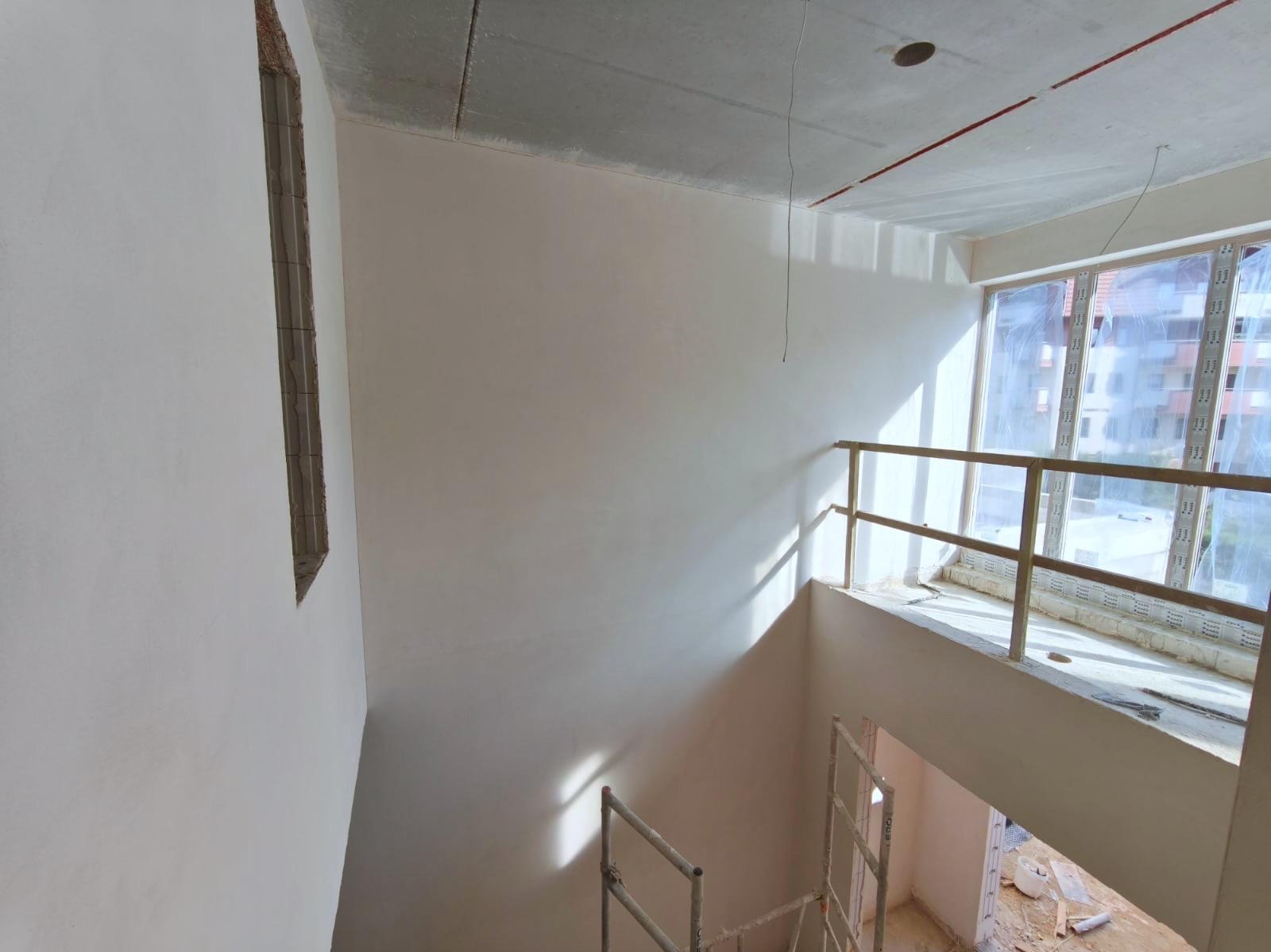 Wnętrze po szpachlowaniu: gładkie, białe ściany, surowy betonowy sufit z instalacjami, okno z widokiem na budynek, fragment rusztowania. Jasne, naturalne światło.