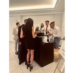 PIOTR NOWAK & CO. EVENTS - Barmani w akcji podczas obsługi gości na eleganckim przyjęciu. Na barze ananasy i akcesoria barmańskie. W tle widoczni goście w strojach wieczorowych.