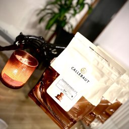 PIOTR NOWAK & CO. EVENTS - Pakiety czekolady Callebaut Fountain Chocolate, świeca zapachowa i dekoracyjna figurka na szklanym stole, sugerujące przygotowania do cateringu.