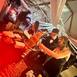 PIOTR NOWAK & CO. EVENTS - Elegancka fontanna czekoladowa na przyjęciu, goście w strojach wieczorowych, dekoracje z balonów i piór, oświetlenie sceniczne, przekąski na stole.