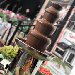 PIOTR NOWAK & CO. EVENTS - Fontanna czekoladowa z owocami (winogrona, truskawki) na stole podczas eventu. Chromowana podstawa, płynąca czekolada. Dekoracja kwiatowa w tle.