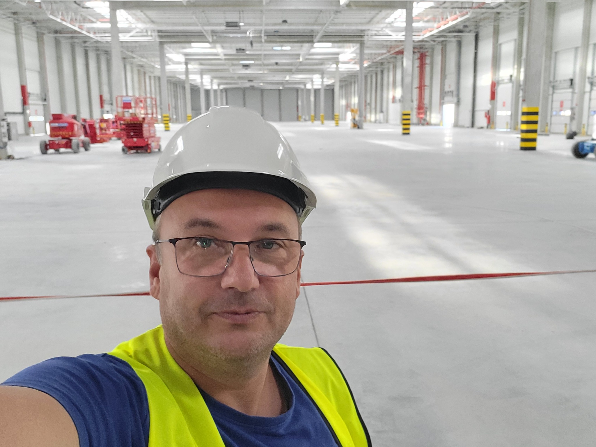 Pracownik w kasku i kamizelce odblaskowej na tle hali magazynowej z podnośnikami, prawdopodobnie po instalacji oświetlenia lub systemu alarmowego.