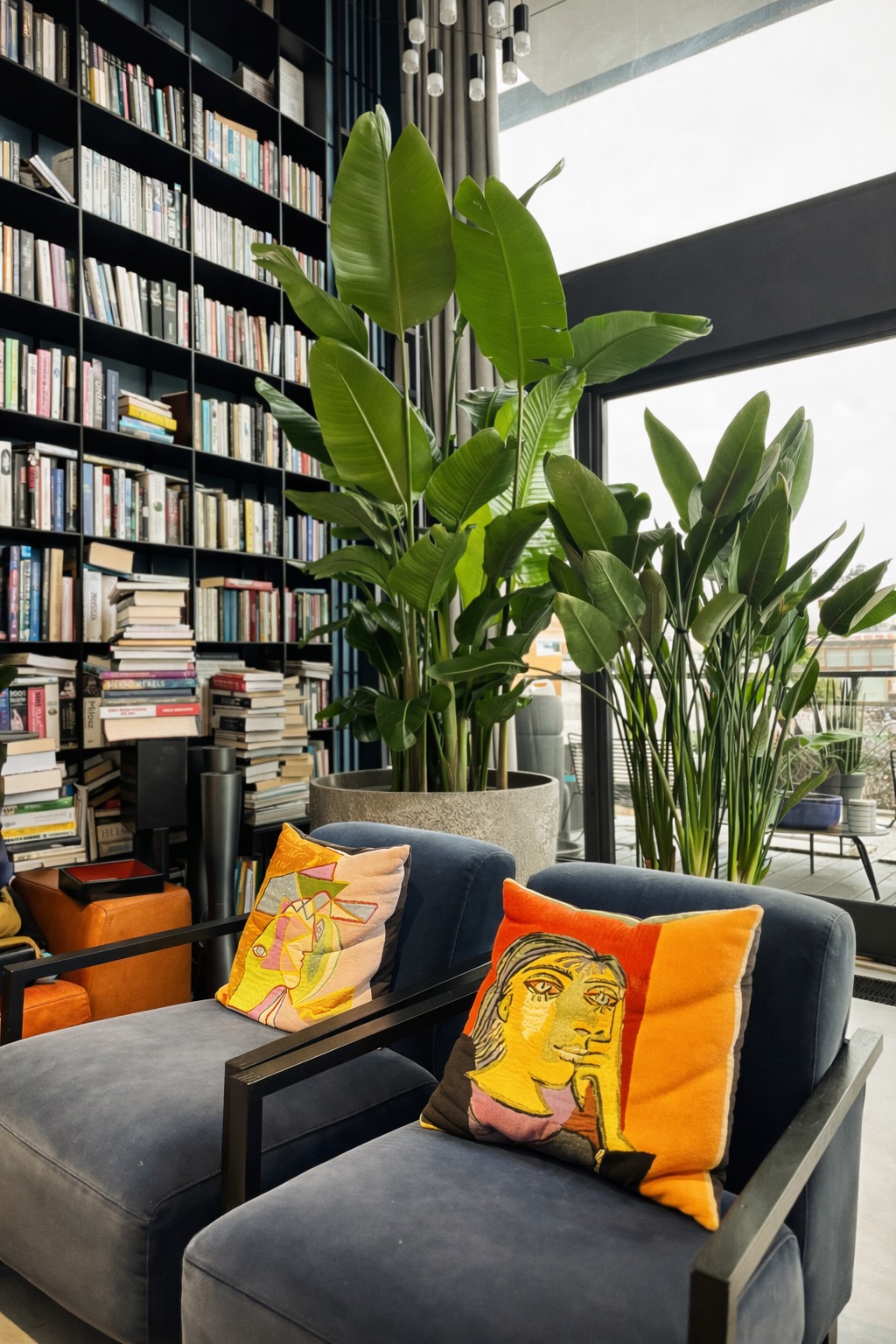 Stylowe wnętrze z biblioteczką, fotelami z artystycznymi poduszkami i dużymi roślinami w donicach przy oknie. Nowoczesny design z elementami sztuki.