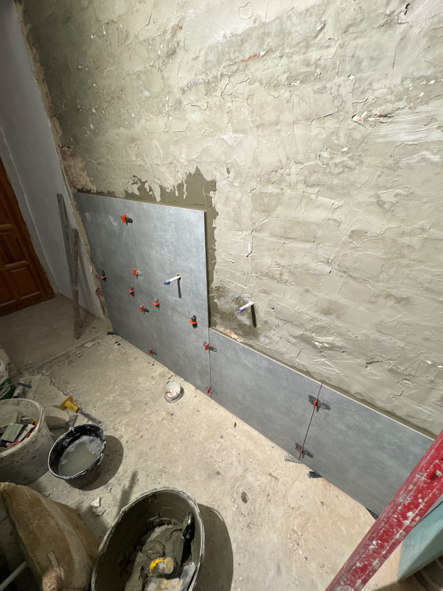 Szare płytki częściowo ułożone na nierównej ścianie z widocznymi dystansami, wiadra z zaprawą i poziomica w tle. Prace glazurnicze w trakcie realizacji.