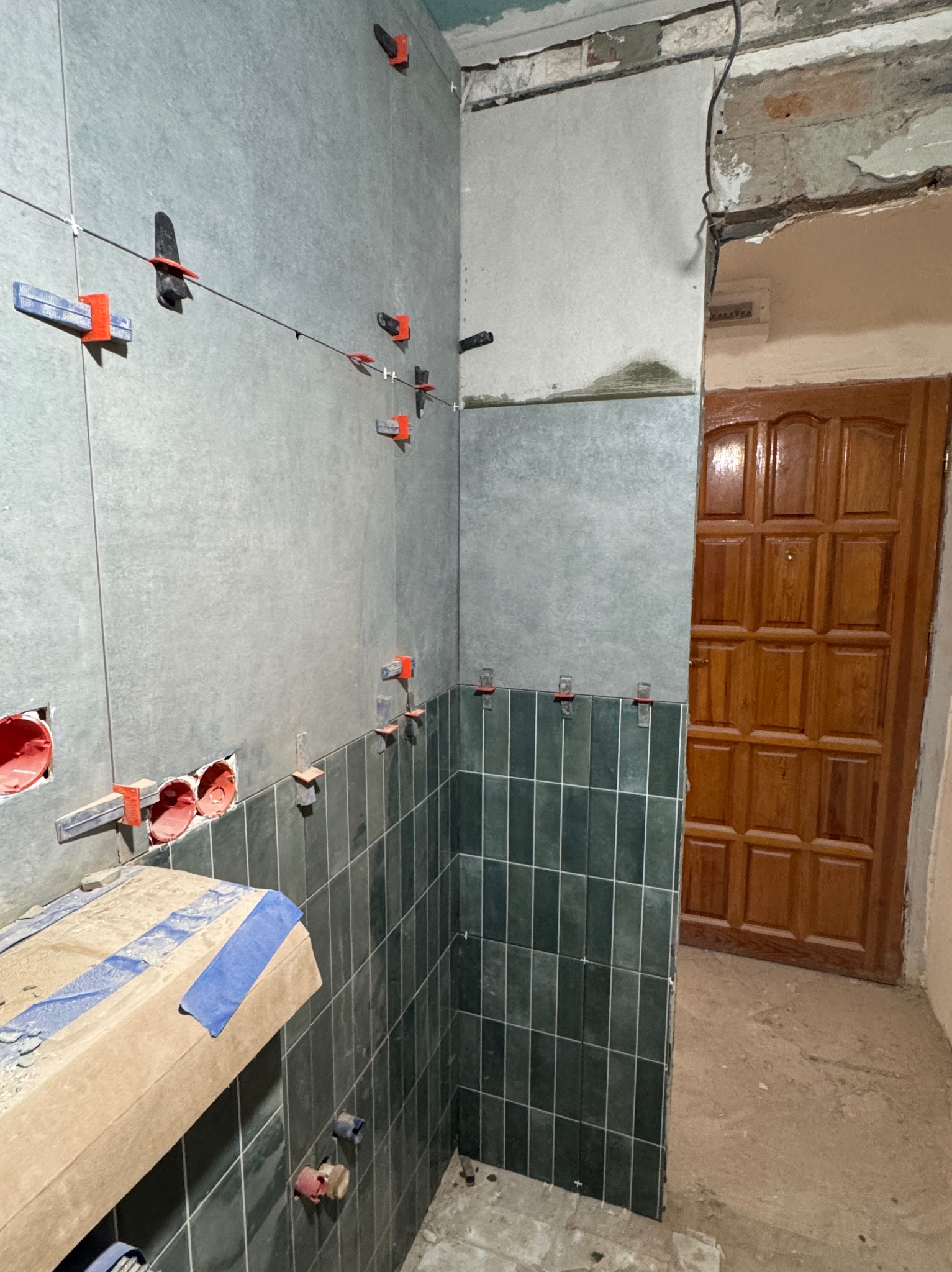 Wnętrze łazienki w trakcie prac glazurniczych: częściowo ułożone ciemnozielone płytki na ścianie, widoczne poziomice i elementy mocujące, surowy stan wykończenia.