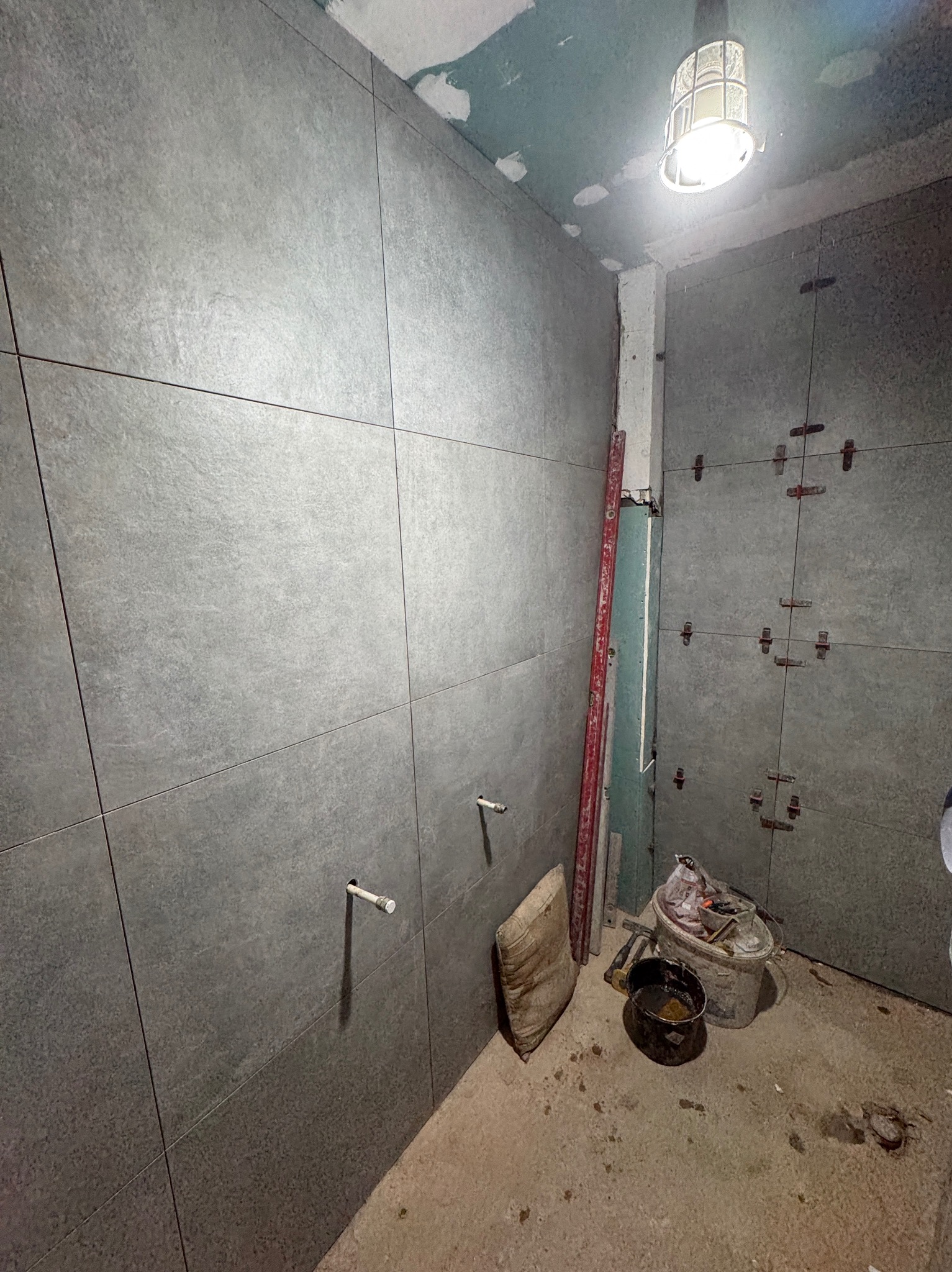 Ściany łazienki wyłożone szarymi, matowymi płytkami wielkoformatowymi. Widoczne narzędzia glazurnicze, poziomica i wystające rurki instalacji wodnej. W tle surowy sufit.