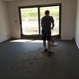 eR-MIX - Pracownik w specjalnych nakładkach wyrównuje świeżo wylaną, szarą posadzkę jastrychową za pomocą zacieraczki mechanicznej w jasnym pomieszczeniu z dużym oknem.