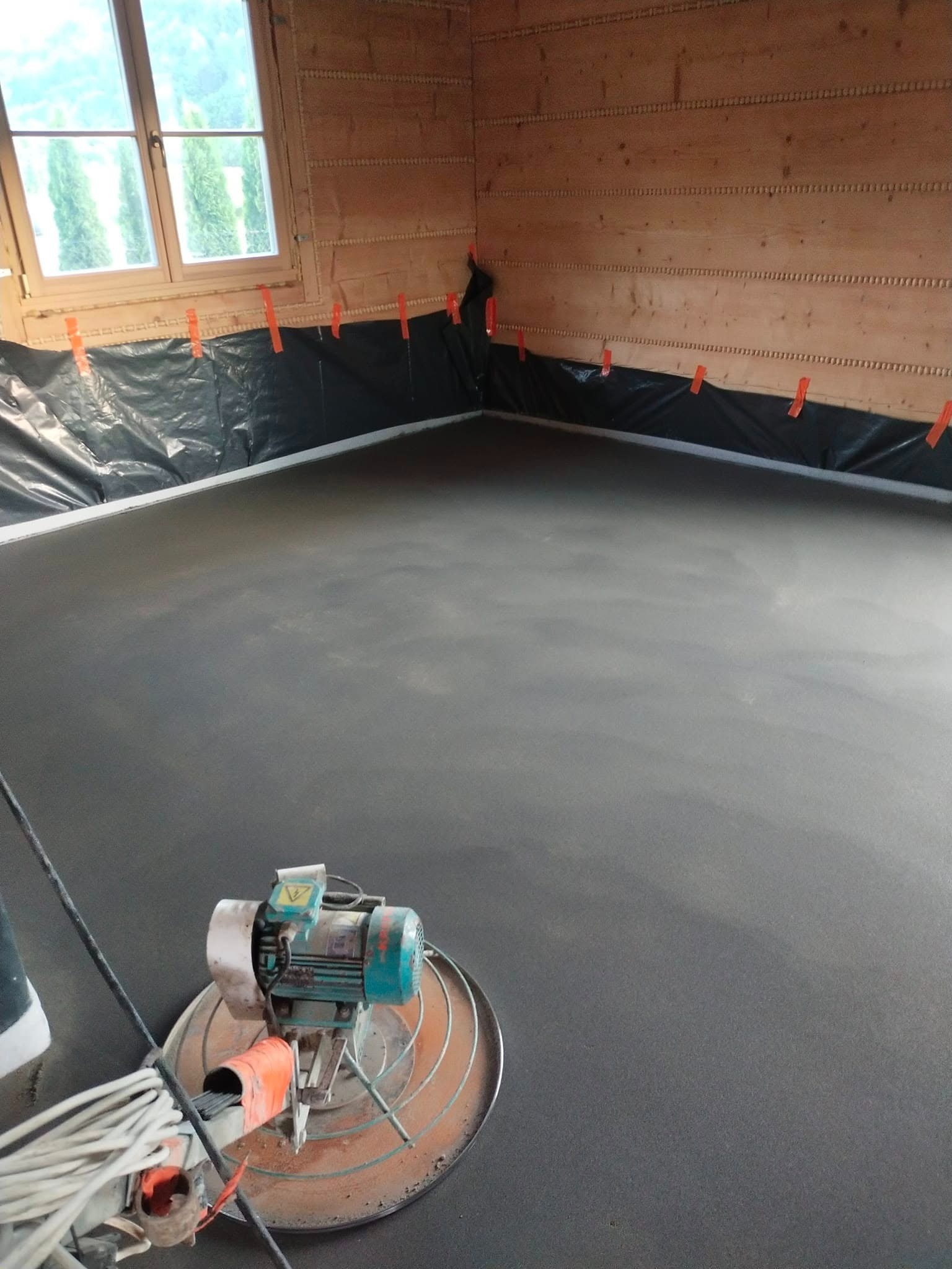 Wyrównana, szara posadzka w drewnianym wnętrzu, zabezpieczona folią przy ścianach. Na pierwszym planie zacieraczka do betonu, gotowa do dalszej obróbki.