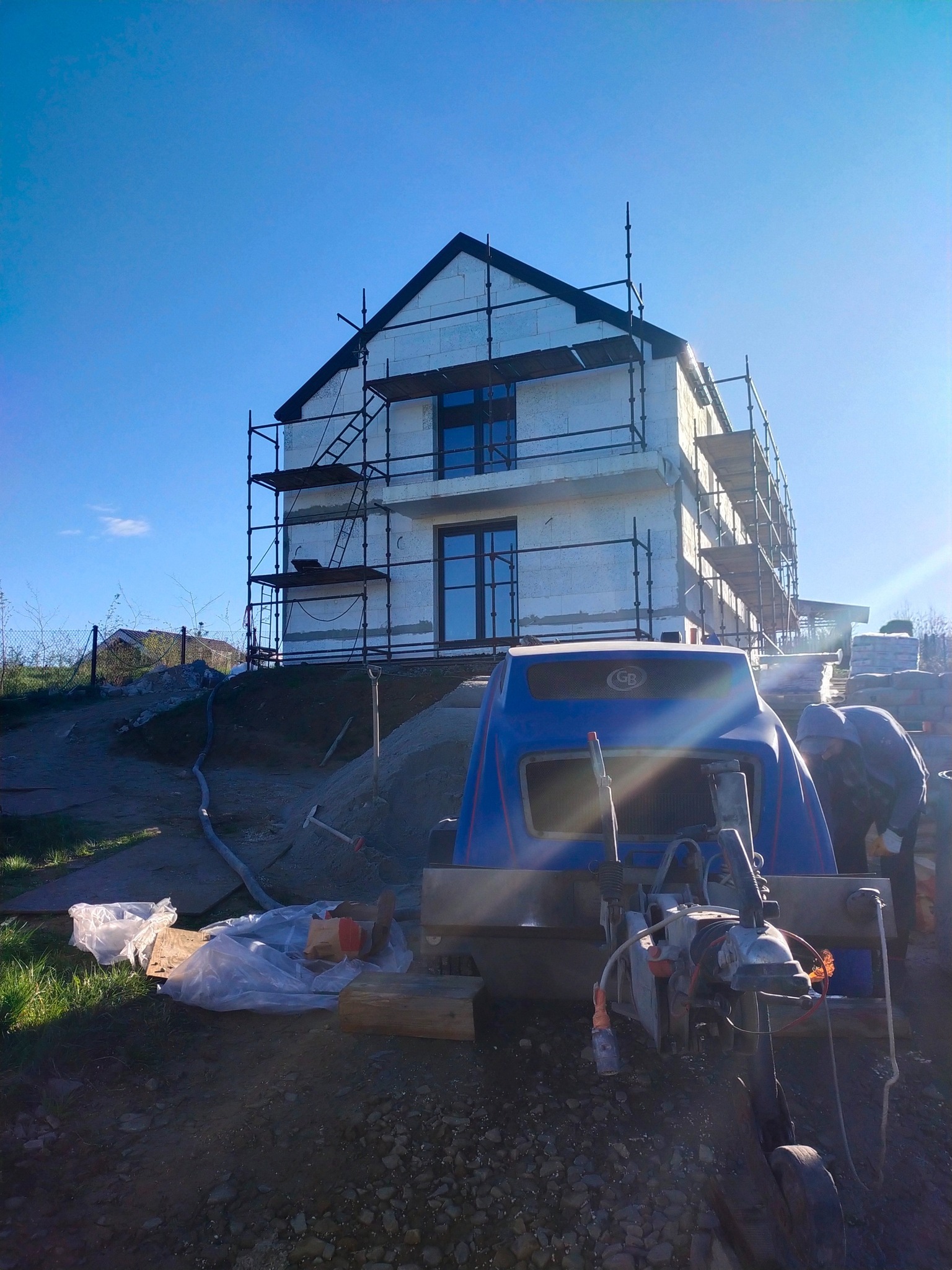 Budowa domu z białych bloczków, rusztowania, niebieska maszyna do wylewek i pracownik w tle, słoneczny dzień.