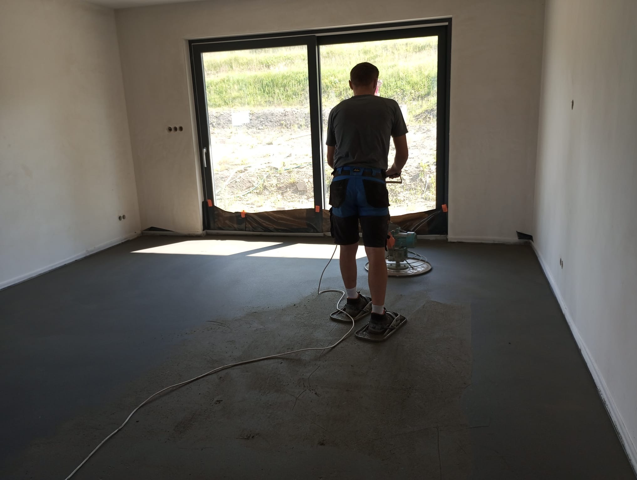 Pracownik w specjalnych nakładkach wyrównuje świeżo wylaną, szarą posadzkę jastrychową za pomocą zacieraczki mechanicznej w jasnym pomieszczeniu z dużym oknem.