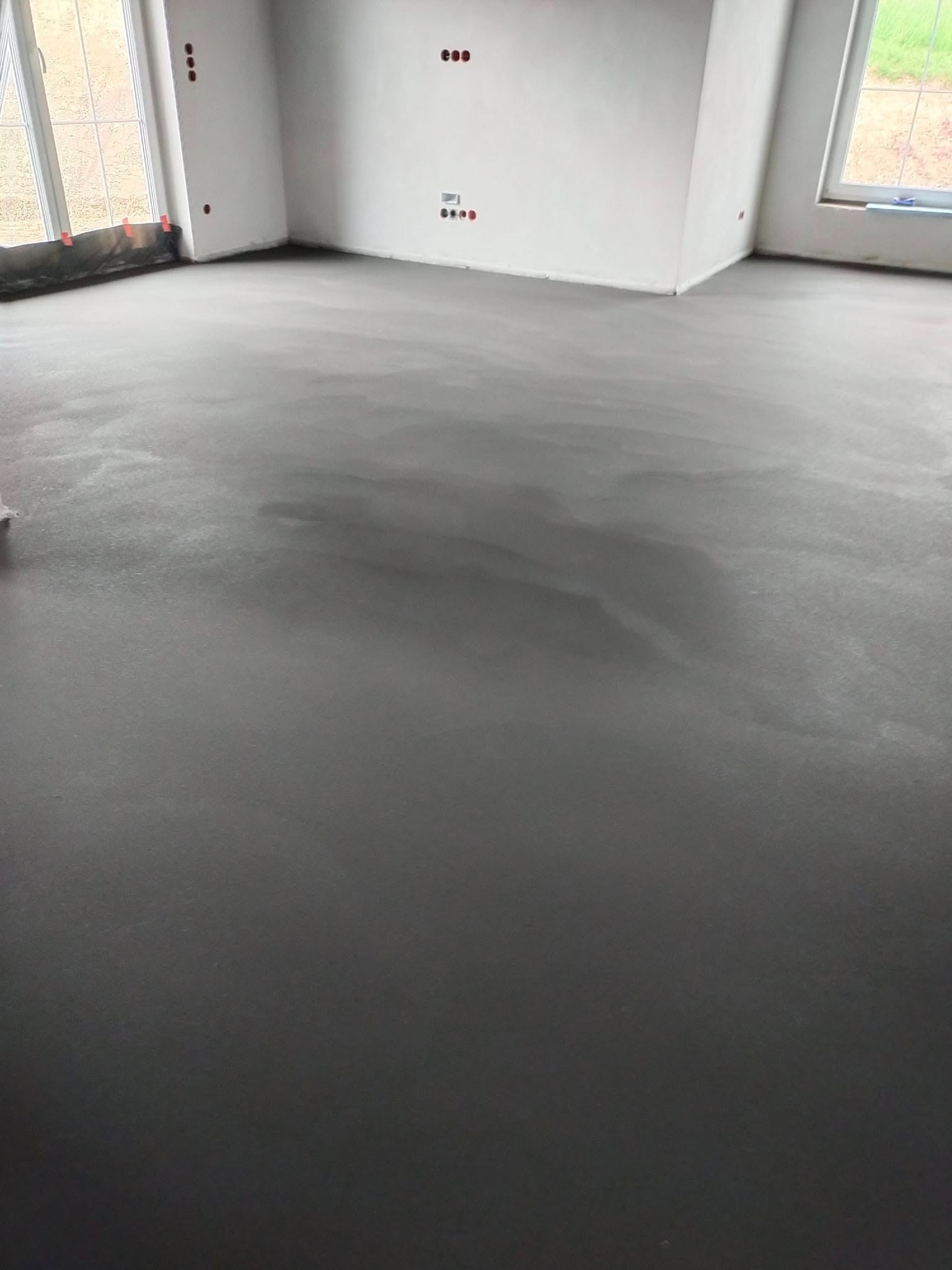 Świeżo wylana, gładka posadzka w jasnym pomieszczeniu z oknami. Widoczne punkty elektryczne w ścianach. Szara, jednolita powierzchnia wylewki.