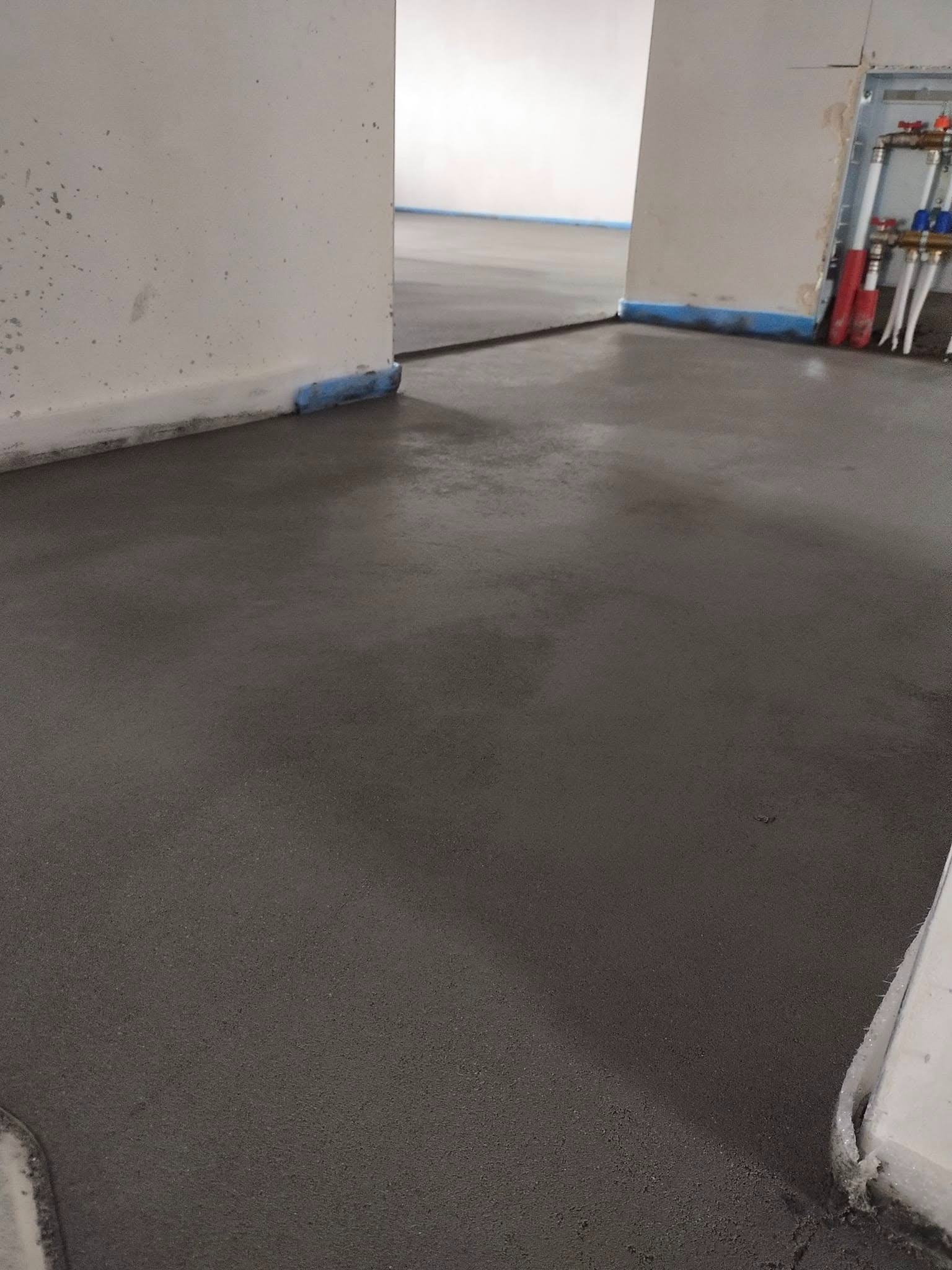 Świeżo wylana, gładka posadzka w szarym odcieniu, zabezpieczona niebieską taśmą przy ścianach. Widoczne rury instalacji grzewczej przy ścianie.